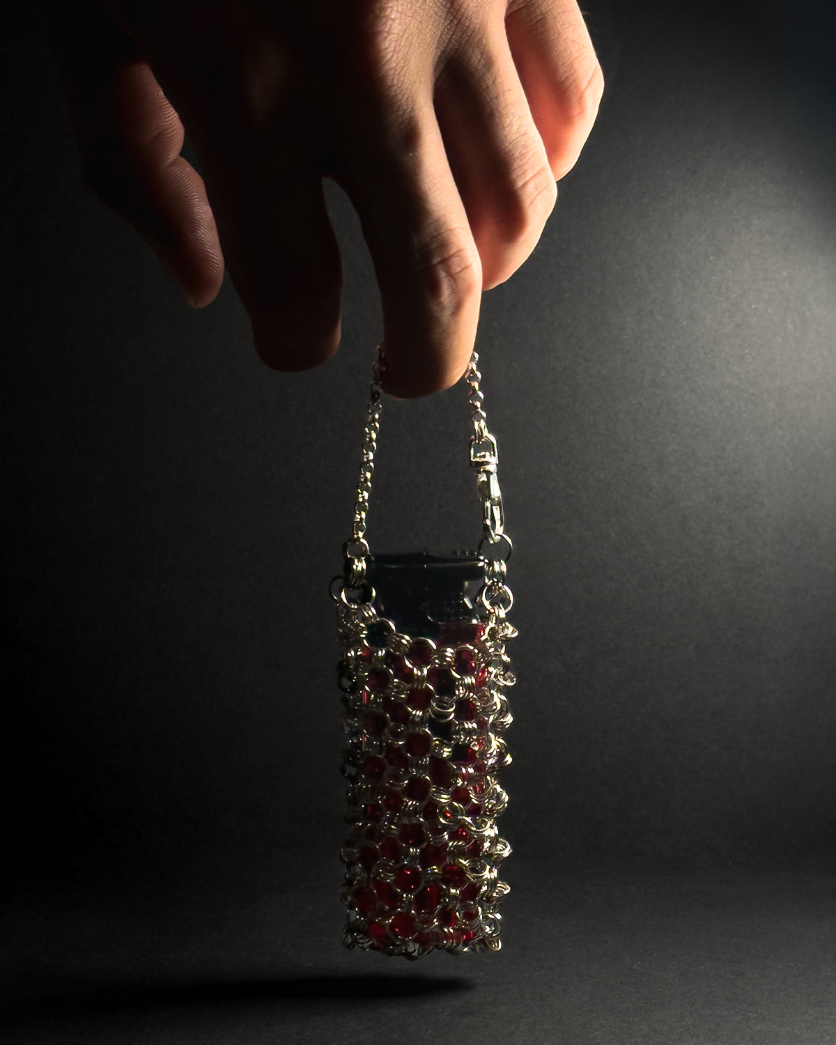 A hand holding a small stainless steel chainmail sleeve filled with lighter against a dark, spotlighted background.