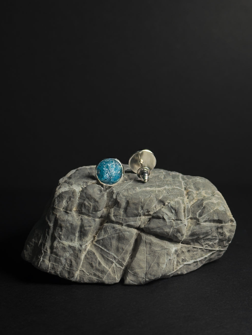 A pair of silver stud earrings, one featuring a round crushed apatite stone inlay and the other showing the silver backing, displayed on a textured gray rock against a solid black background.