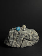 A pair of silver stud earrings, one featuring a round crushed apatite stone inlay and the other showing the silver backing, displayed on a textured gray rock against a solid black background.