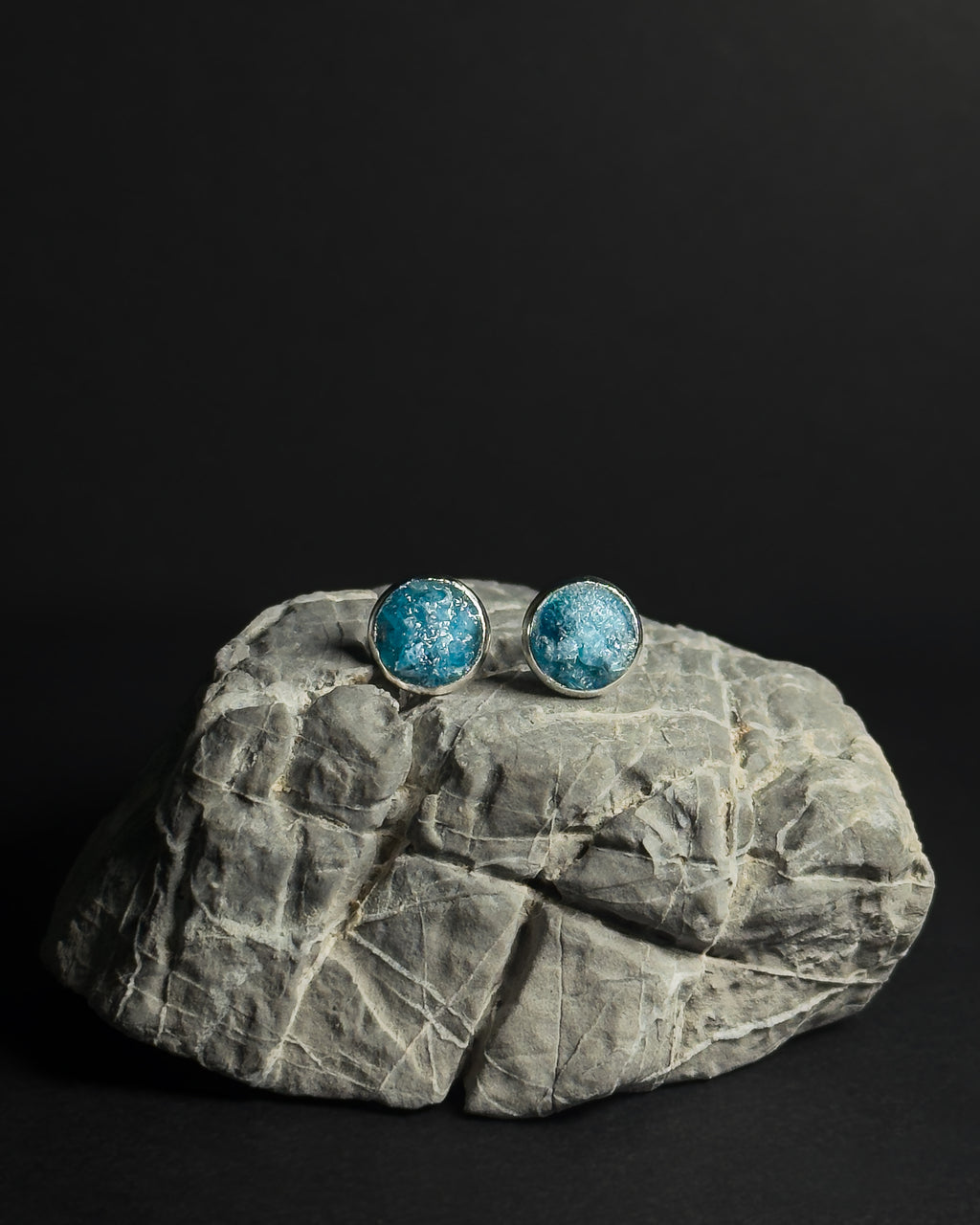 A pair of round stud earrings with crushed apatite stone inlay in silver bezel settings, displayed on a gray stone prop against a solid black background.