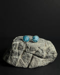A pair of round stud earrings with crushed apatite stone inlay in silver bezel settings, displayed on a gray stone prop against a solid black background.