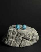 A pair of round stud earrings with crushed apatite stone inlay in silver bezel settings, displayed on a gray stone prop against a solid black background.