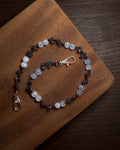 A beaded phone charm keychian with light blue and dark blue faceted beads, accented with silver-toned stainless steel lobster clasp ends, displayed on a wooden board over a dark wood surface.