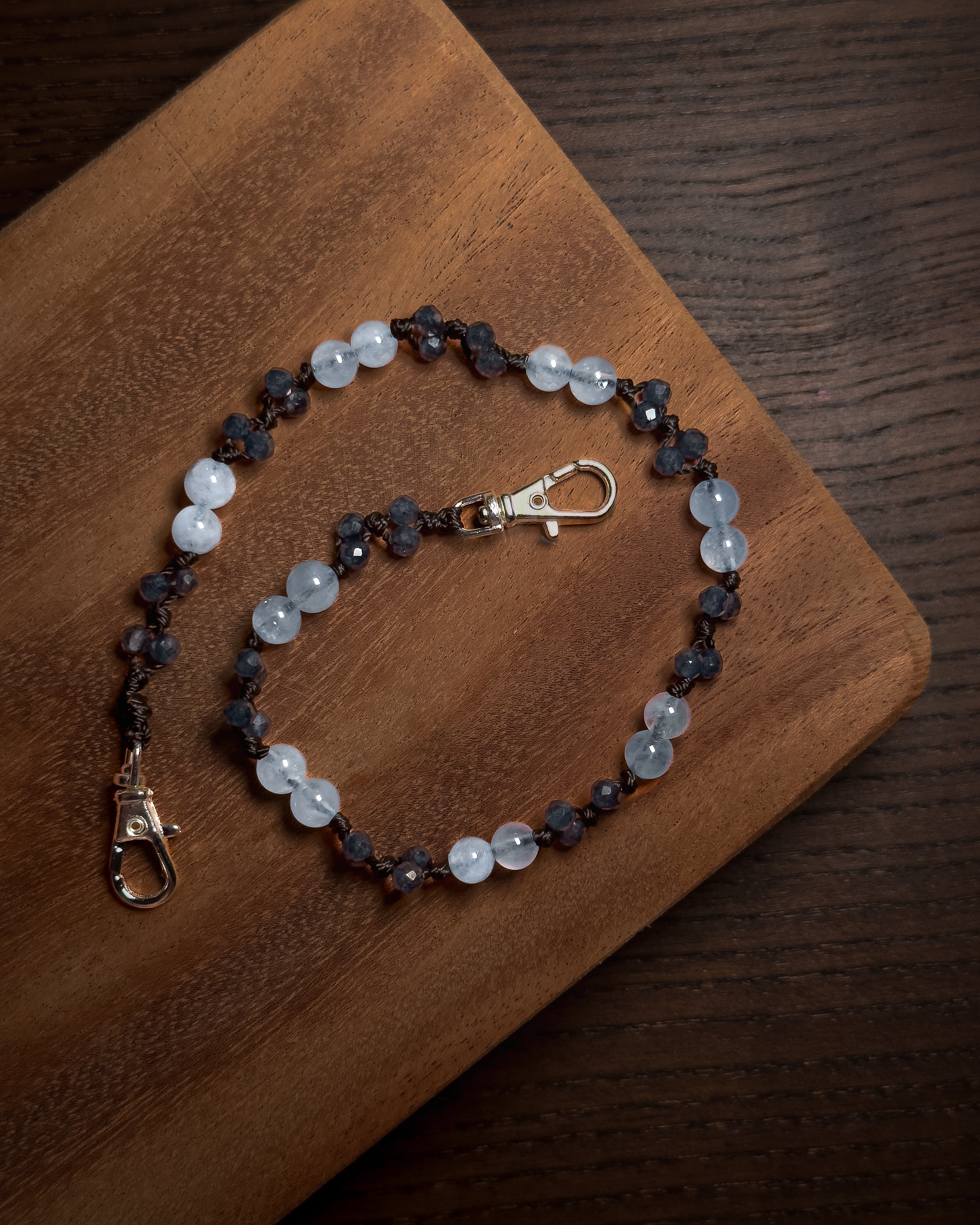 A beaded phone charm keychian with light blue and dark blue faceted beads, accented with silver-toned stainless steel lobster clasp ends, displayed on a wooden board over a dark wood surface.