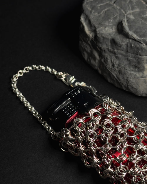 A close-up of a silver chainmail-style sleeve with lighter, attached to a silver chain strap. A dark textured stone prop sits in the background on a black surface.
