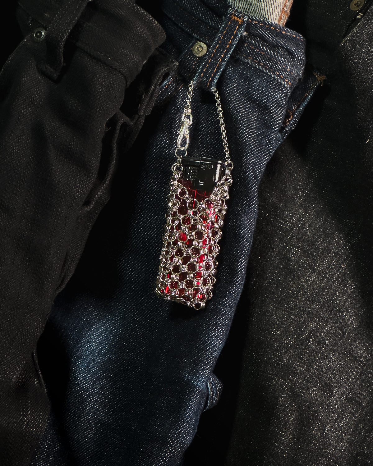 A close-up of a stainless steel chainmail sleeve with lighter, hanging from the waistband of dark blue denim jeans.