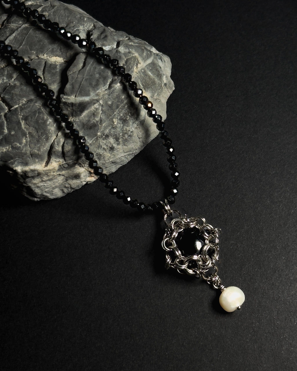 Stainless steel chainmail necklace with 14mm black onyx pendant, freshwater pearl and faceted spinel beads on a natural textured grey rock against a dark background.
