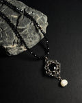 Stainless steel chainmail necklace with 14mm black onyx pendant, freshwater pearl and faceted spinel beads on a natural textured grey rock against a dark background.