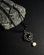 Stainless steel chainmail necklace with 14mm black onyx pendant, freshwater pearl and faceted spinel beads on a natural textured grey rock against a dark background.