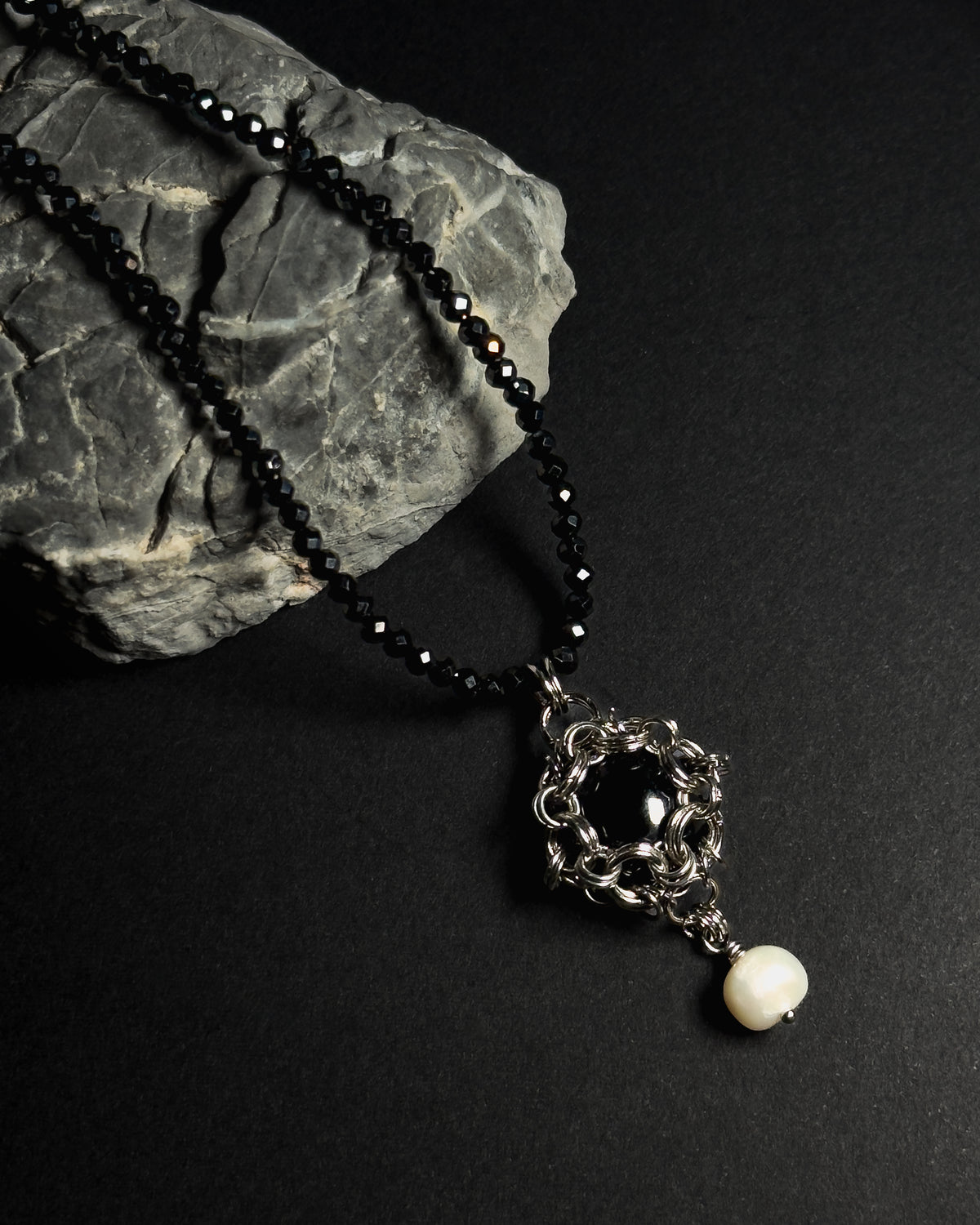 Stainless steel chainmail necklace with 14mm black onyx pendant, freshwater pearl and faceted spinel beads on a natural textured grey rock against a dark background.