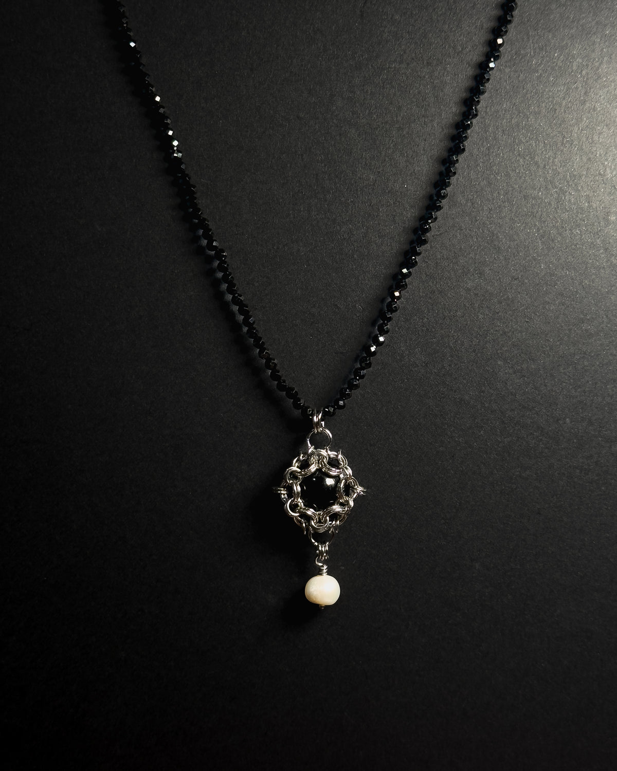 Stainless steel chainmail necklace with 14mm black onyx pendant, freshwater pearl and faceted spinel beads against a dark background.