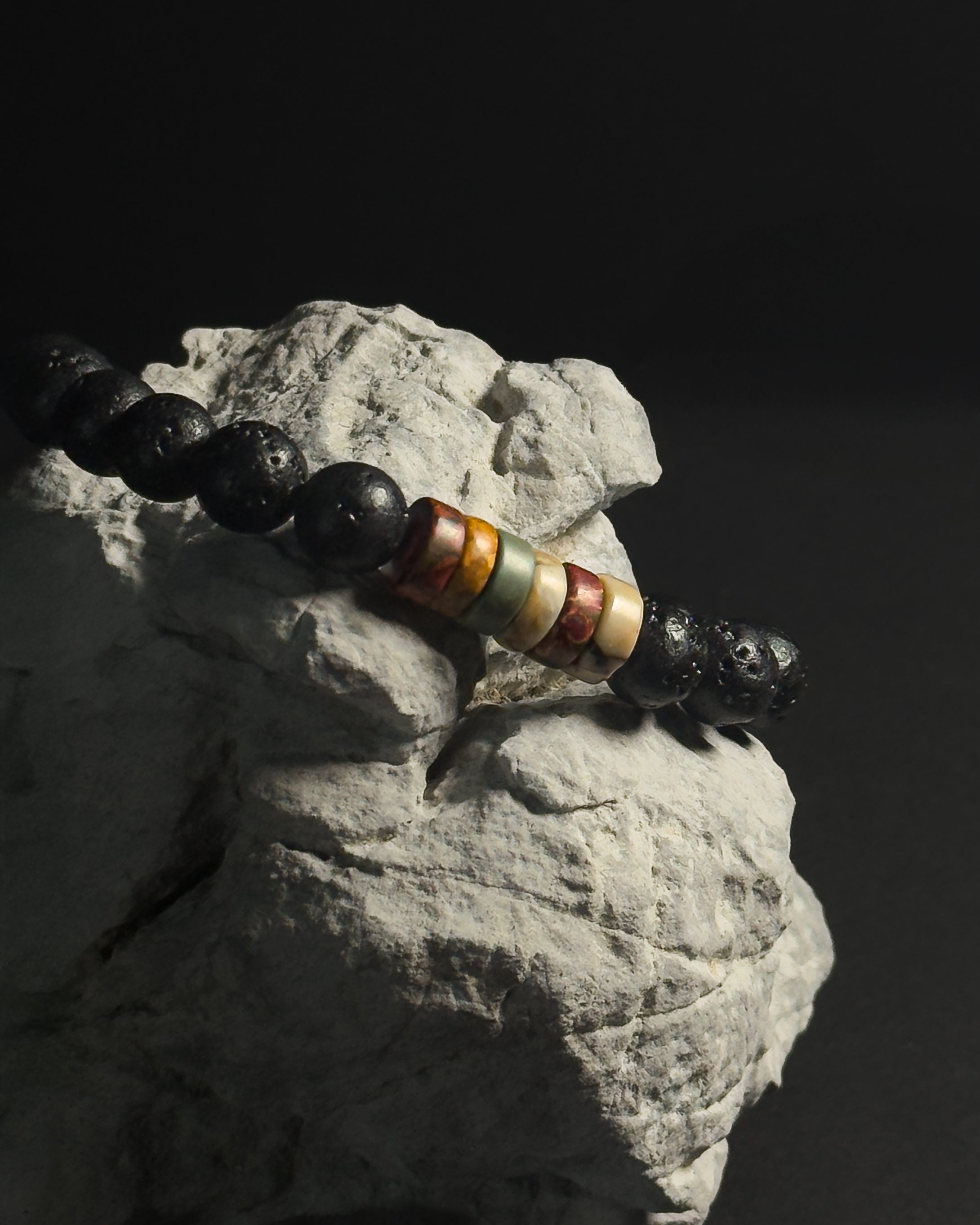 A beaded bracelet with glossy black round beads and a central cluster of striped cylindrical beads in orange, cream, and brown, displayed on a textured white rock against a dark background.
