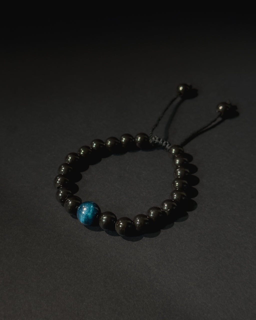 Black beaded bracelet with a blue bead on a dark background. Black beaded bracelet with blue tiger eye.