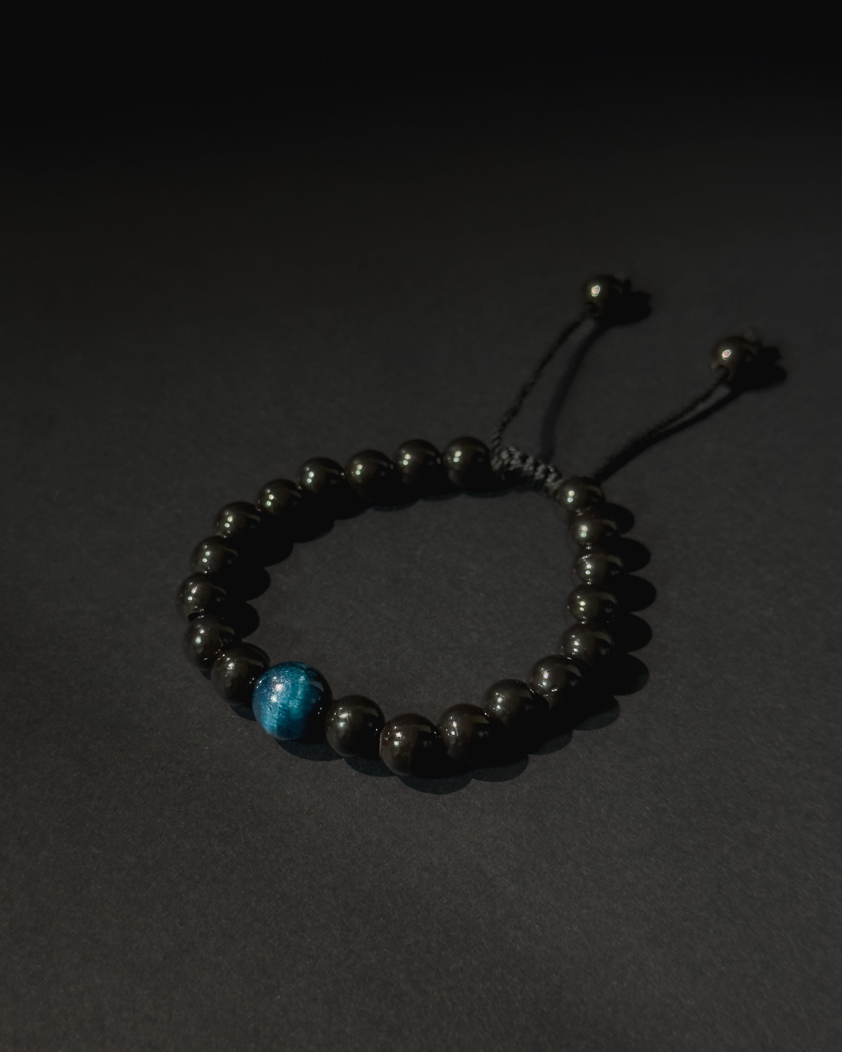 Black beaded bracelet with a blue bead on a dark background. Black beaded bracelet with blue tiger eye.
