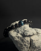 Bracelet with black and blue beads on a rock against a dark background. Black beaded bracelet with blue tiger eye.