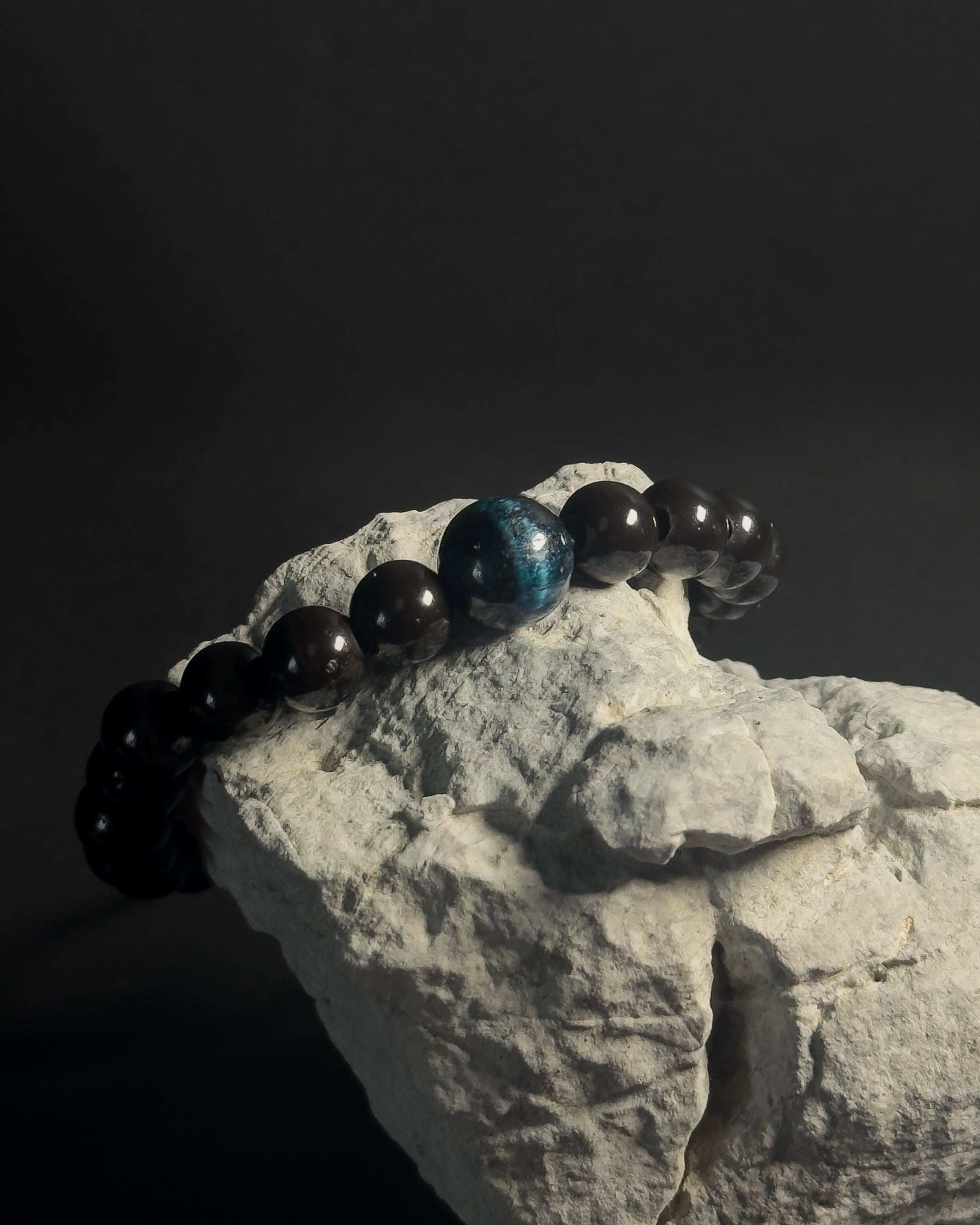 Bracelet with black and blue beads on a rock against a dark background. Black beaded bracelet with blue tiger eye.