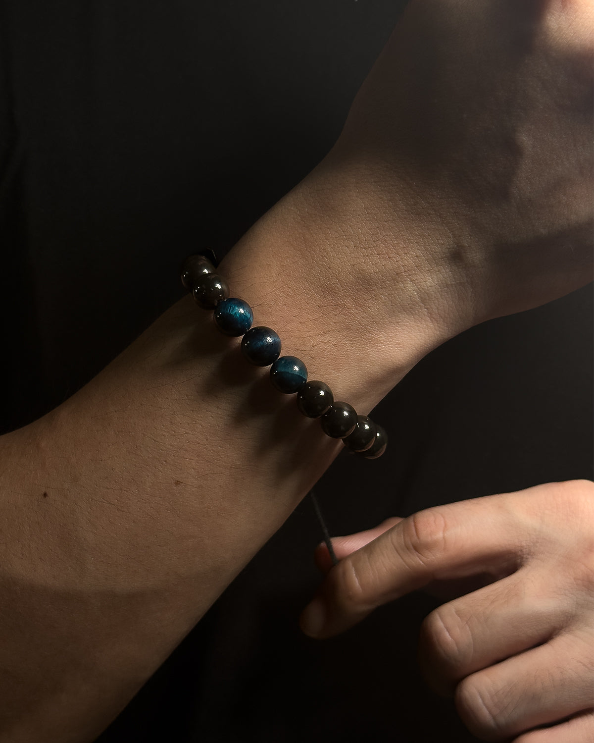 Close-up of a wrist wearing a black beaded bracelet with blue tiger eye against a dark background