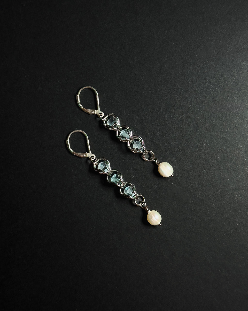 A pair of silver drop earrings featuring light blue topaz beads, silver wirework, and white pearl, displayed on a dark textured background.