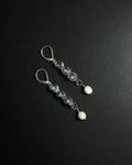 A pair of silver drop earrings featuring light blue topaz beads, silver wirework, and white pearl, displayed on a dark textured background.