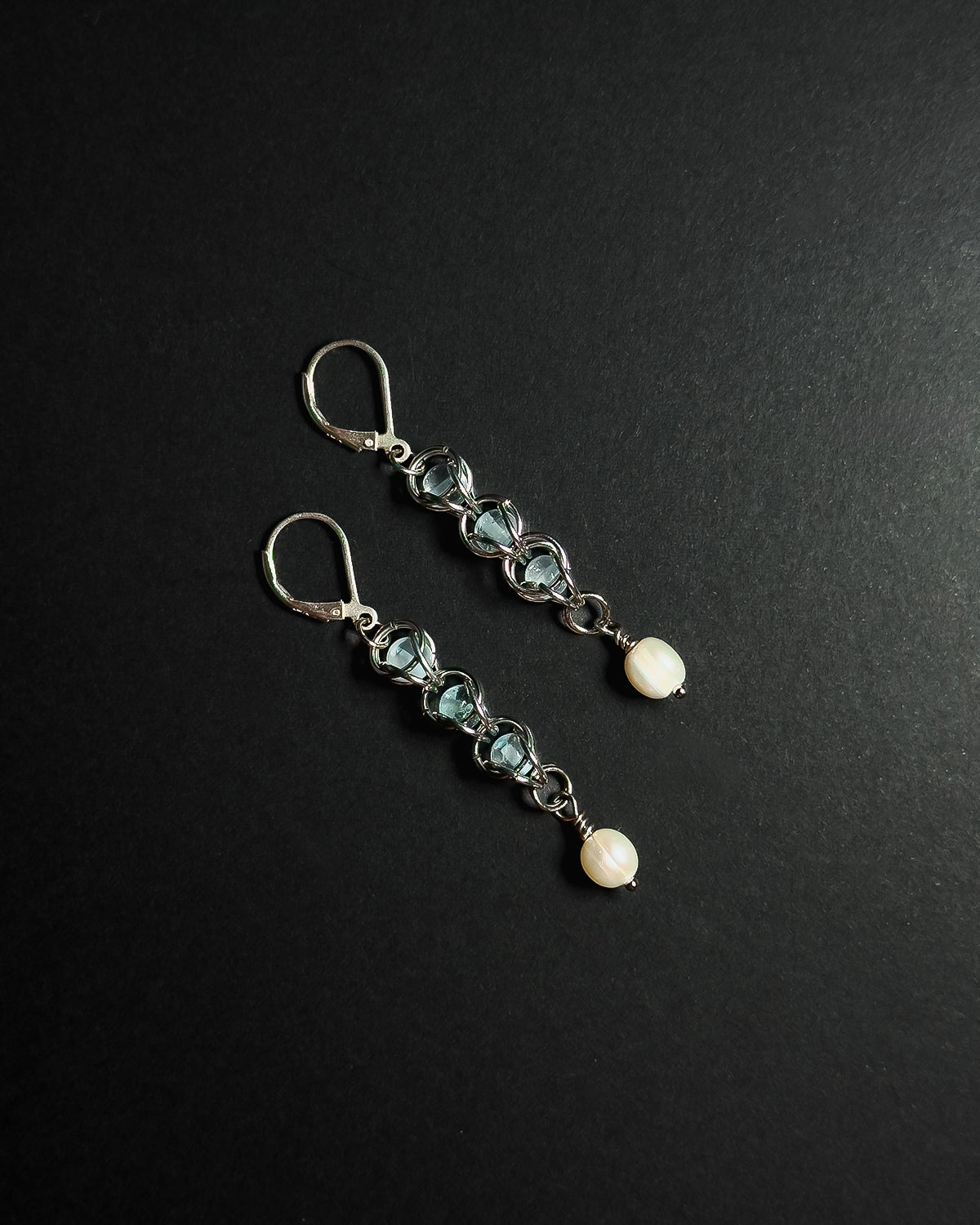 A pair of silver drop earrings featuring light blue topaz beads, silver wirework, and white pearl, displayed on a dark textured background.