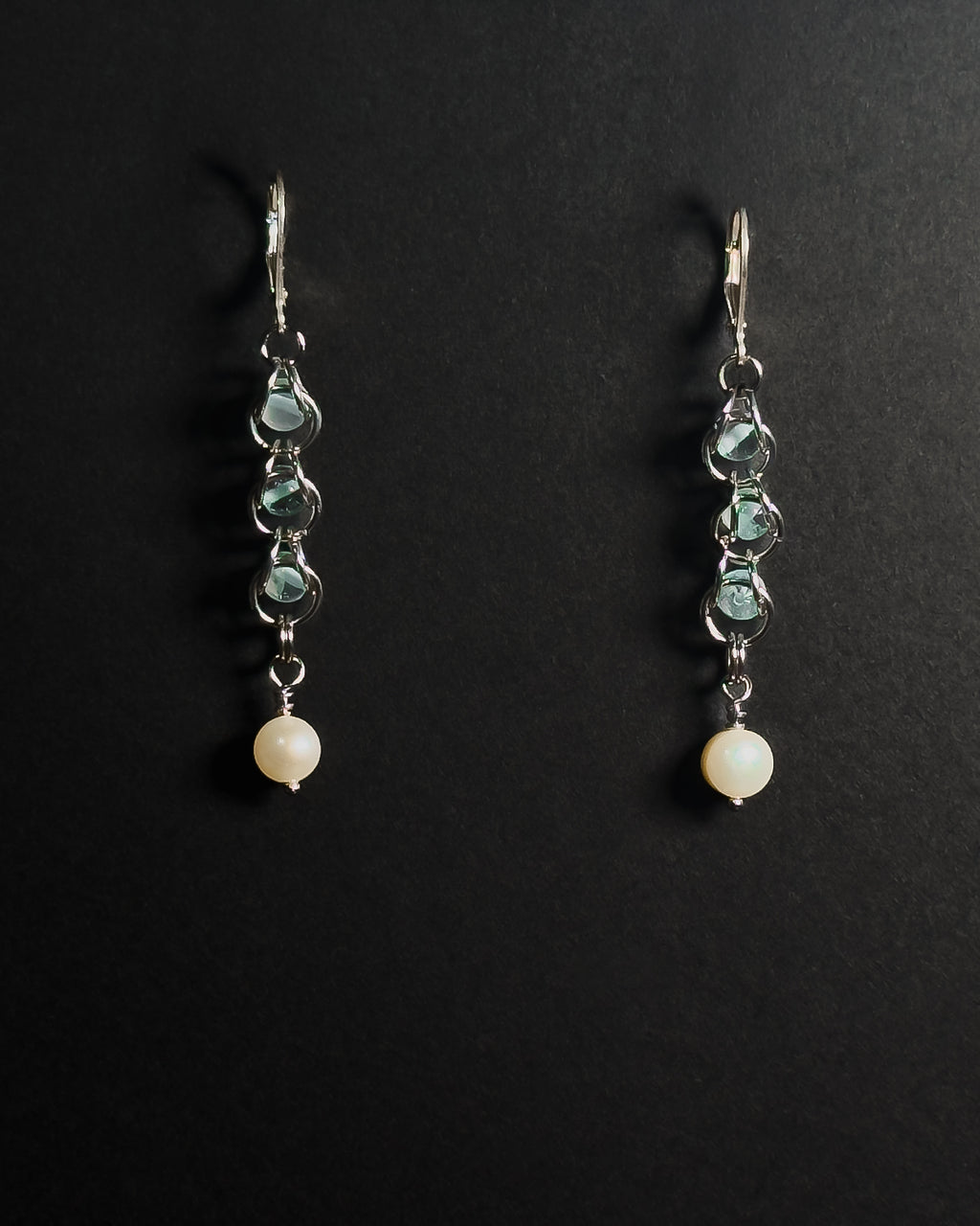 A pair of silver leverback dangle earrings featuring blue topaz capture bead and a round white pearl at the bottom, displayed on a dark background.