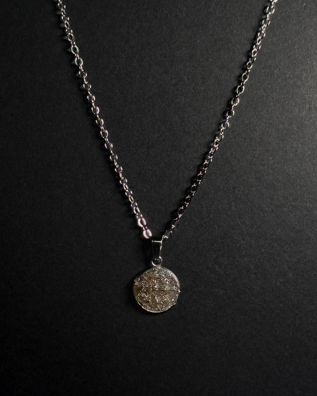 A silver-toned chain necklace with a round textured crushed smokey quartz stone pendant, displayed against a dark black background.