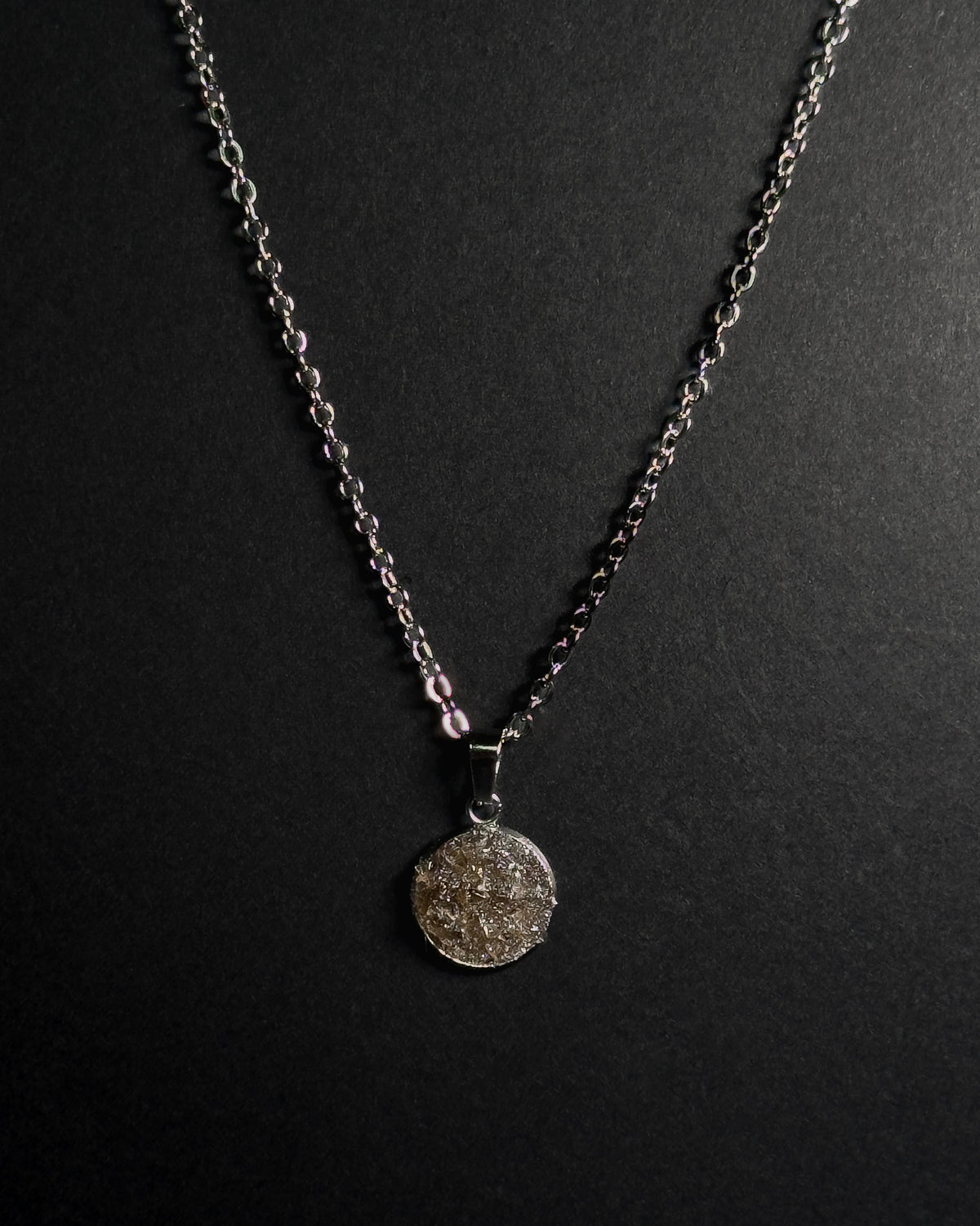 A silver-toned chain necklace with a round textured crushed smokey quartz stone pendant, displayed against a dark black background.