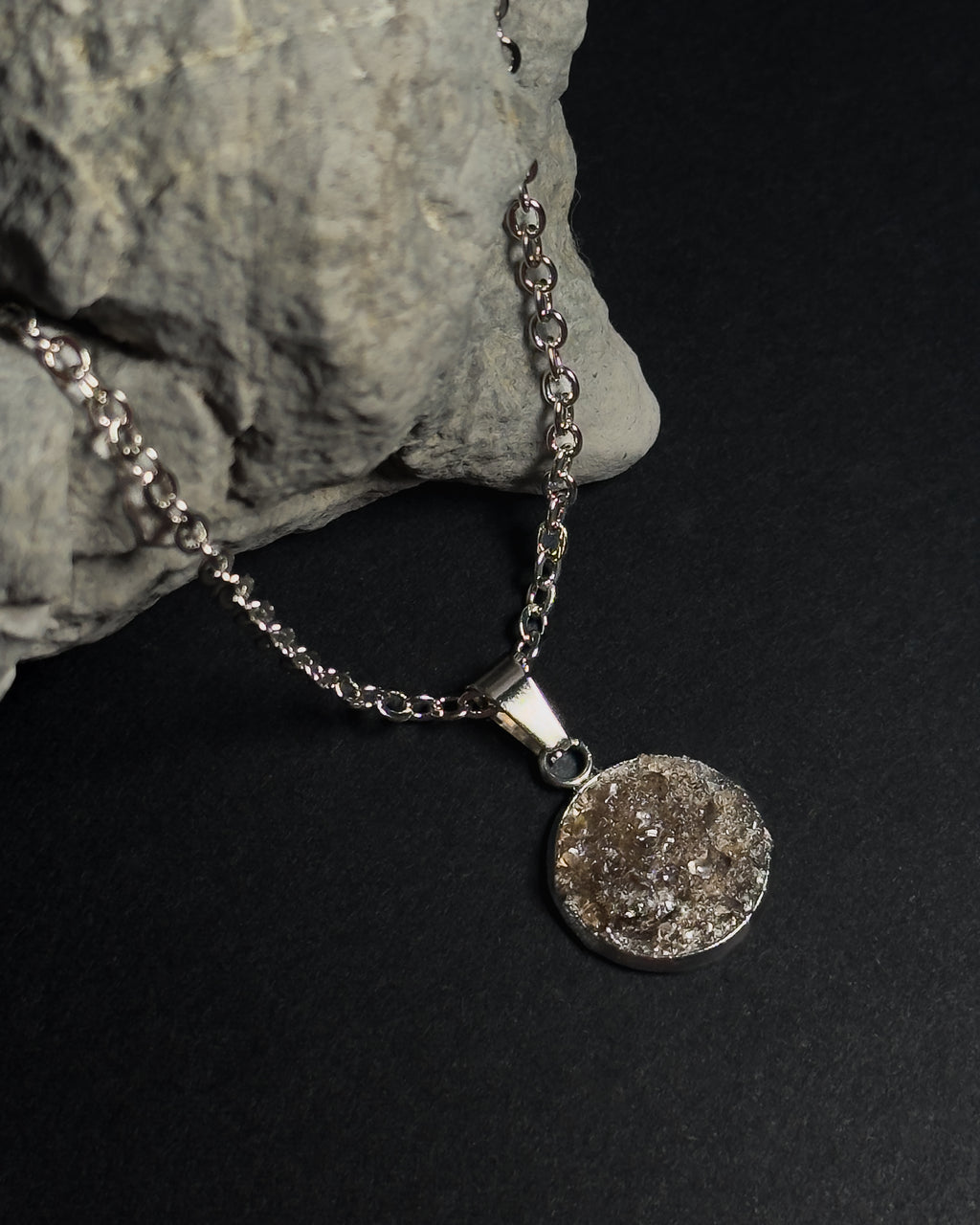 A silver chain necklace with a round, textured crushed smokey quartz inlay pendant, displayed on a dark background next to a gray rock prop.