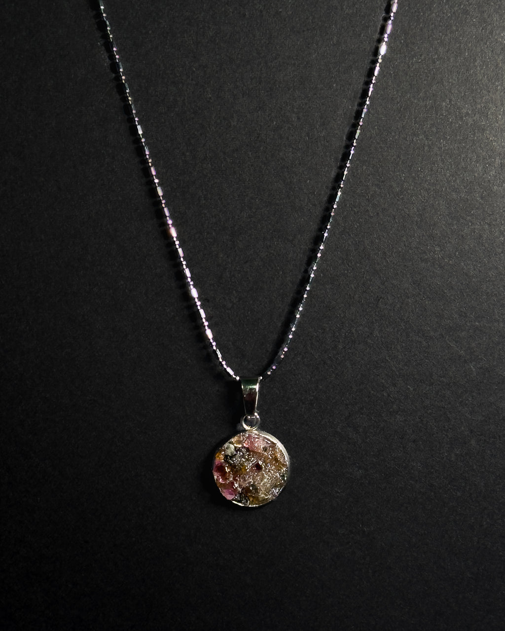 A silver beaded chain necklace with a round pendant containing crushed rainbow tourmaline, displayed against a dark textured background.