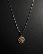 A silver beaded chain necklace with a round pendant containing crushed rainbow tourmaline, displayed against a dark textured background.