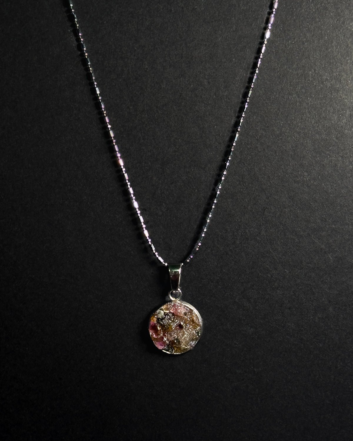 A silver beaded chain necklace with a round pendant containing crushed rainbow tourmaline, displayed against a dark textured background.