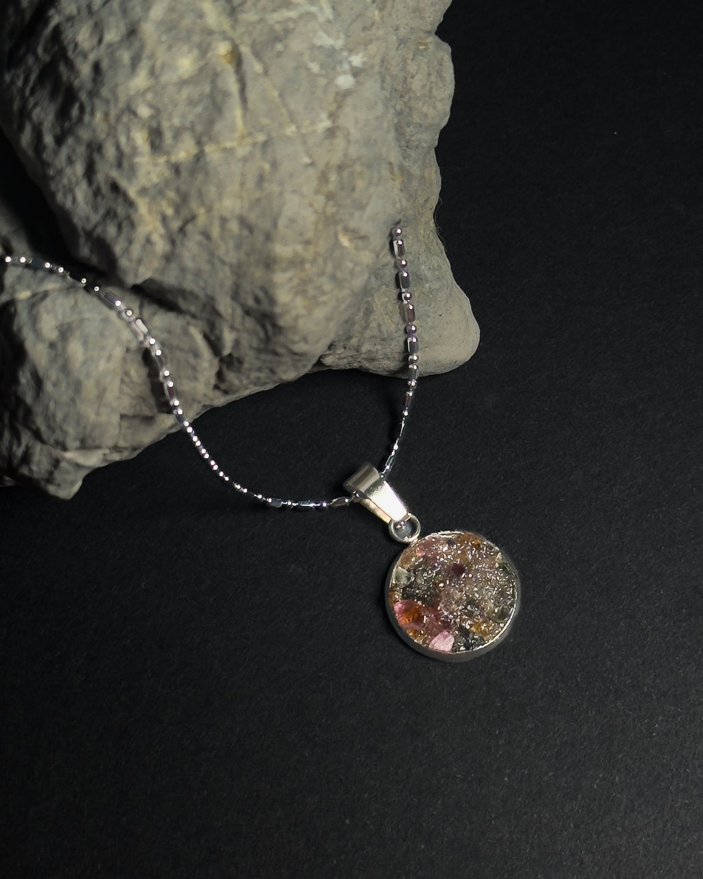 A silver beaded chain necklace with a round pendant containing crushed rainbow tourmaline stone, displayed on a dark background next to a textured gray rock.