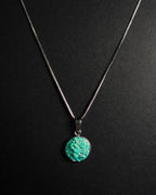 A silver chain necklace with a round, crudhed turquoise pendant on a dark black background.
