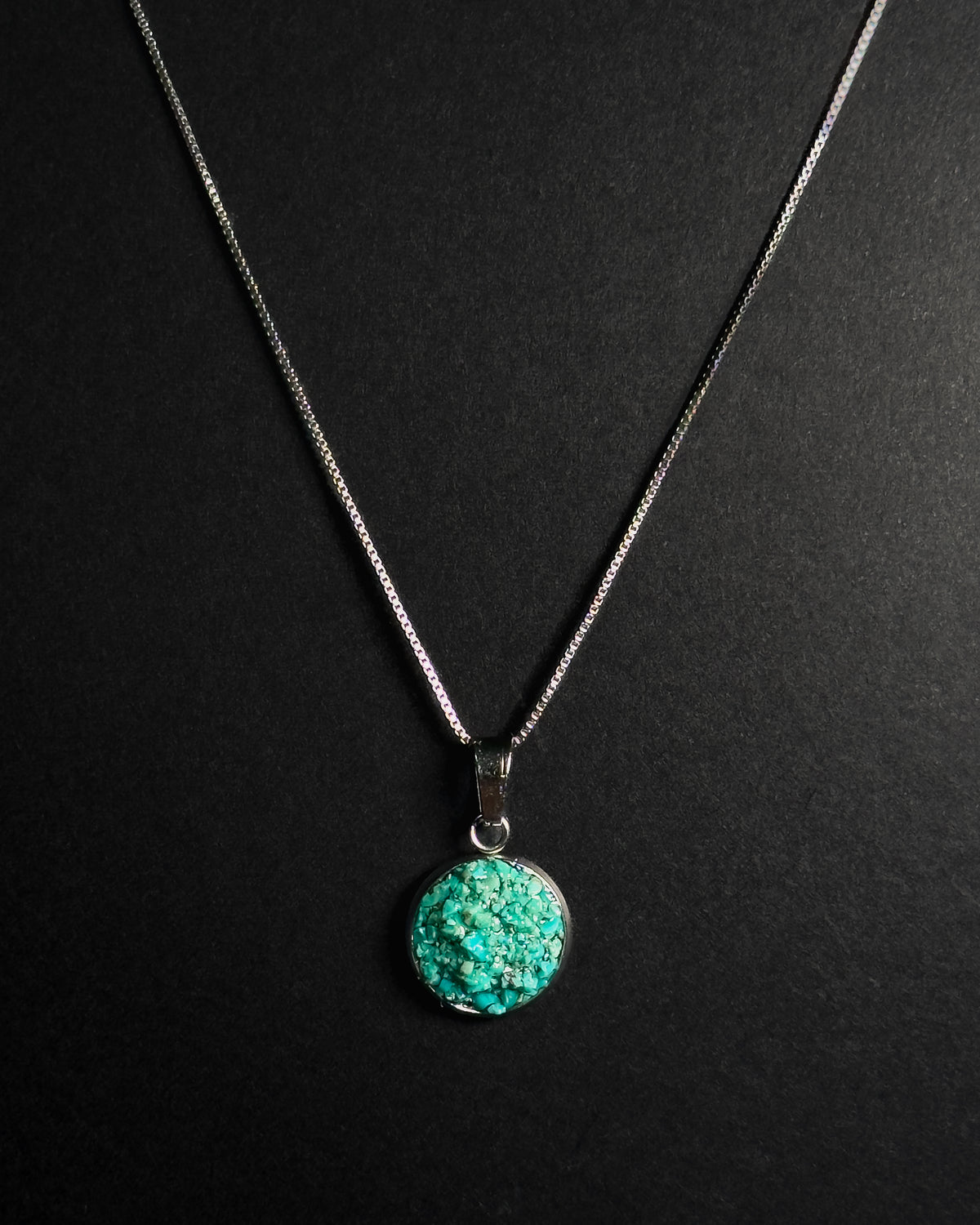 A silver chain necklace with a round, crudhed turquoise pendant on a dark black background.