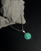 A silver chain necklace with a round turquoise pendant, displayed on a dark background next to a gray rock.