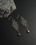A pair of silver-toned chainmail dangle earrings with a dangle freshwater pearl drop, displayed on a dark textured surface next to a gray stone.