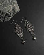 A pair of silver-toned chainmail dangle earrings with a dangle freshwater pearl drop, displayed on a dark textured surface next to a gray stone.