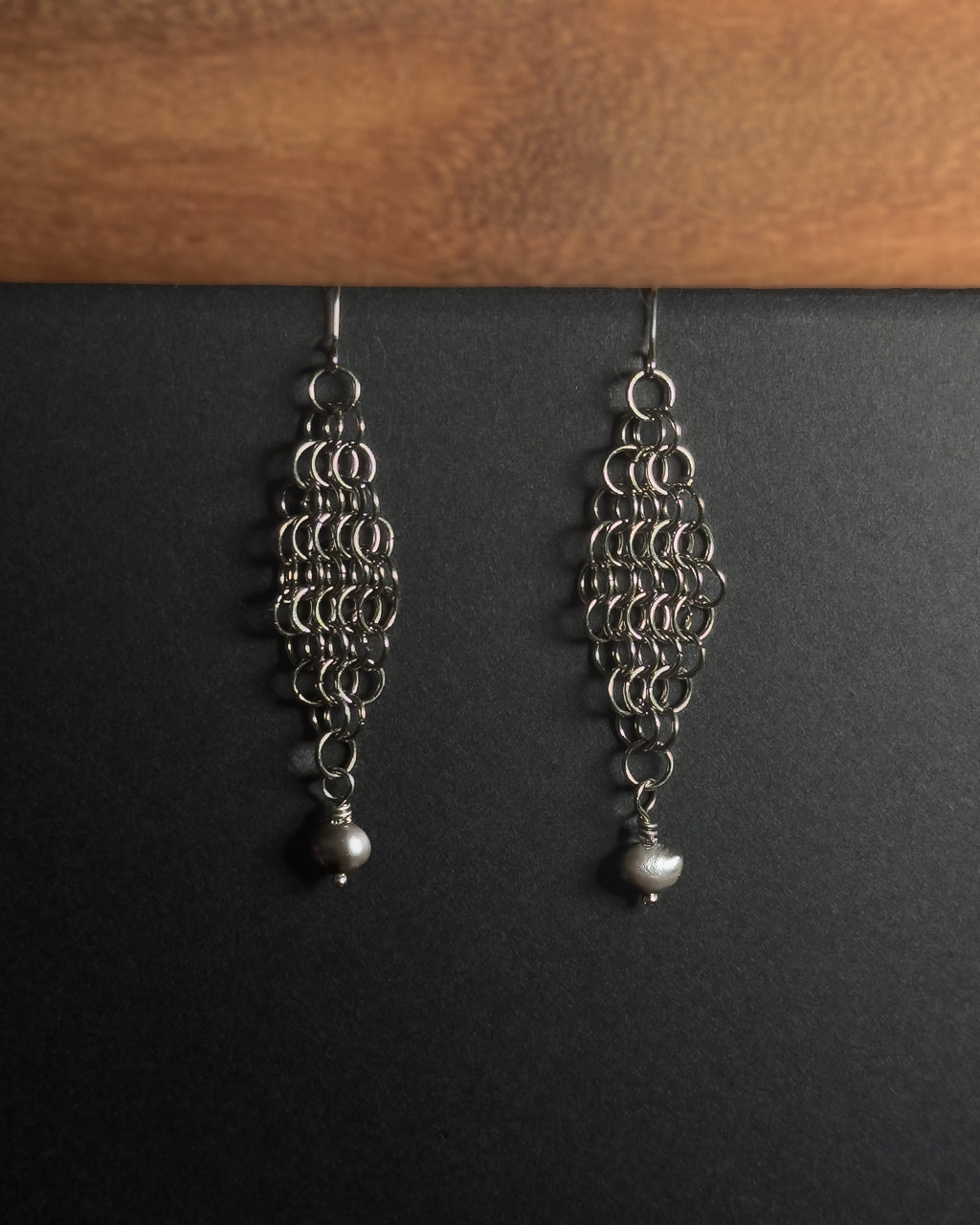 A pair of silver-toned dangle earrings with intricate circular link patterns and small pearl drops, displayed against a dark background with a wooden surface at the top.