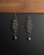 A pair of silver-toned dangle earrings with intricate circular link patterns and small pearl drops, displayed against a dark background with a wooden surface at the top.
