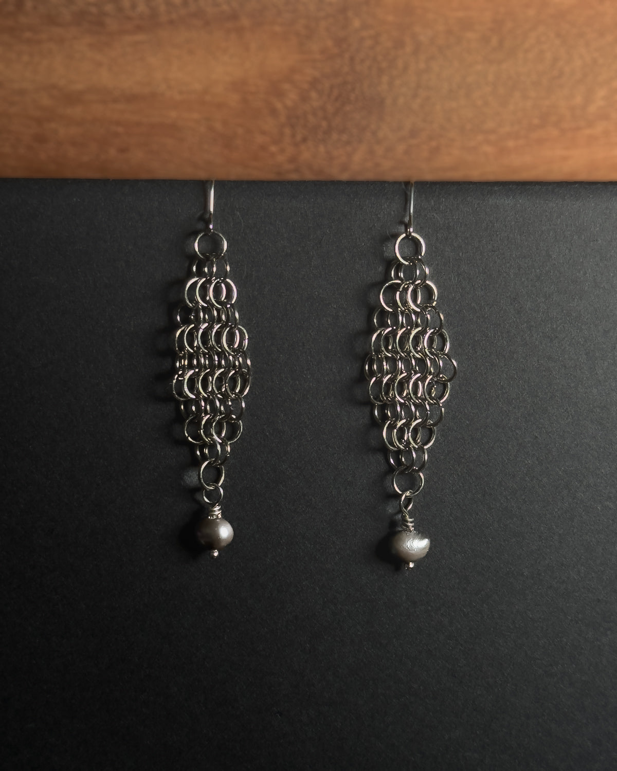 A pair of silver-toned dangle earrings with intricate circular link patterns and small pearl drops, displayed against a dark background with a wooden surface at the top.