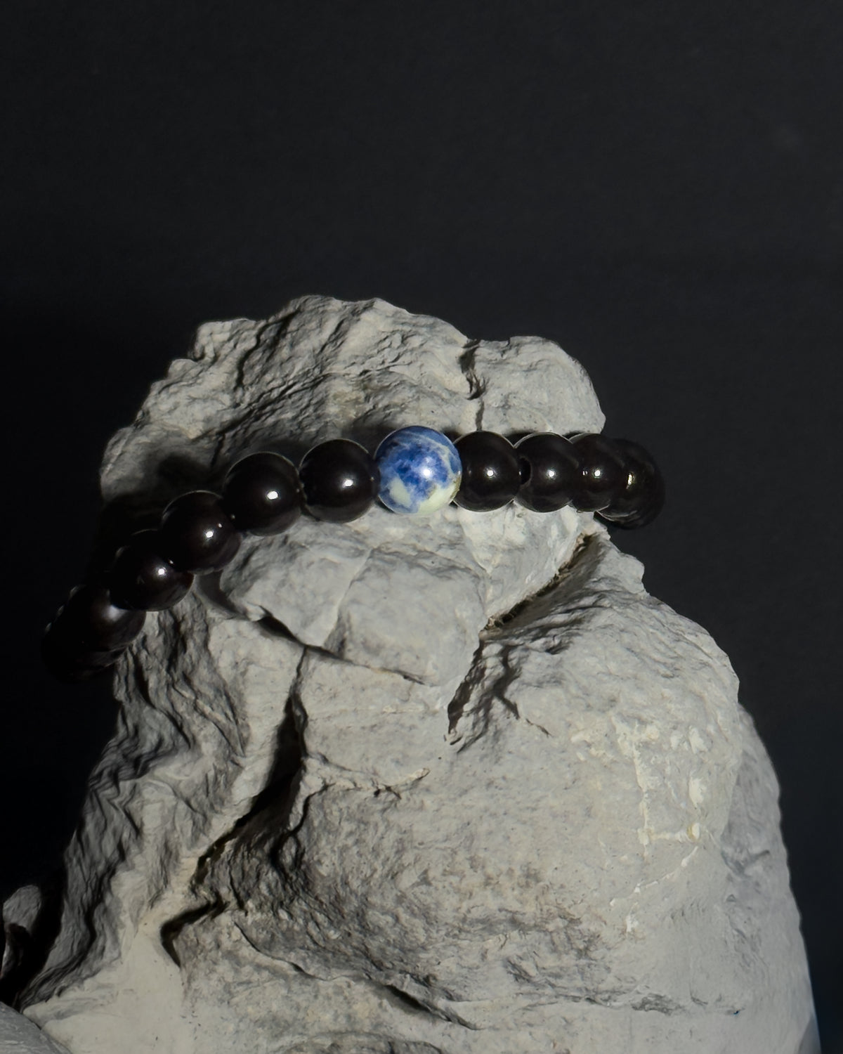 A beaded bracelet with mostly tourmaline and a sodalite bead, displayed on a gray rock against a black background.
