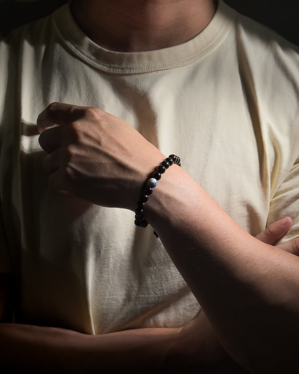 A close-up of a person wearing a black tourmaline beaded bracelet with a sodalite center stone on their wrist, wearing a beige t-shirt.