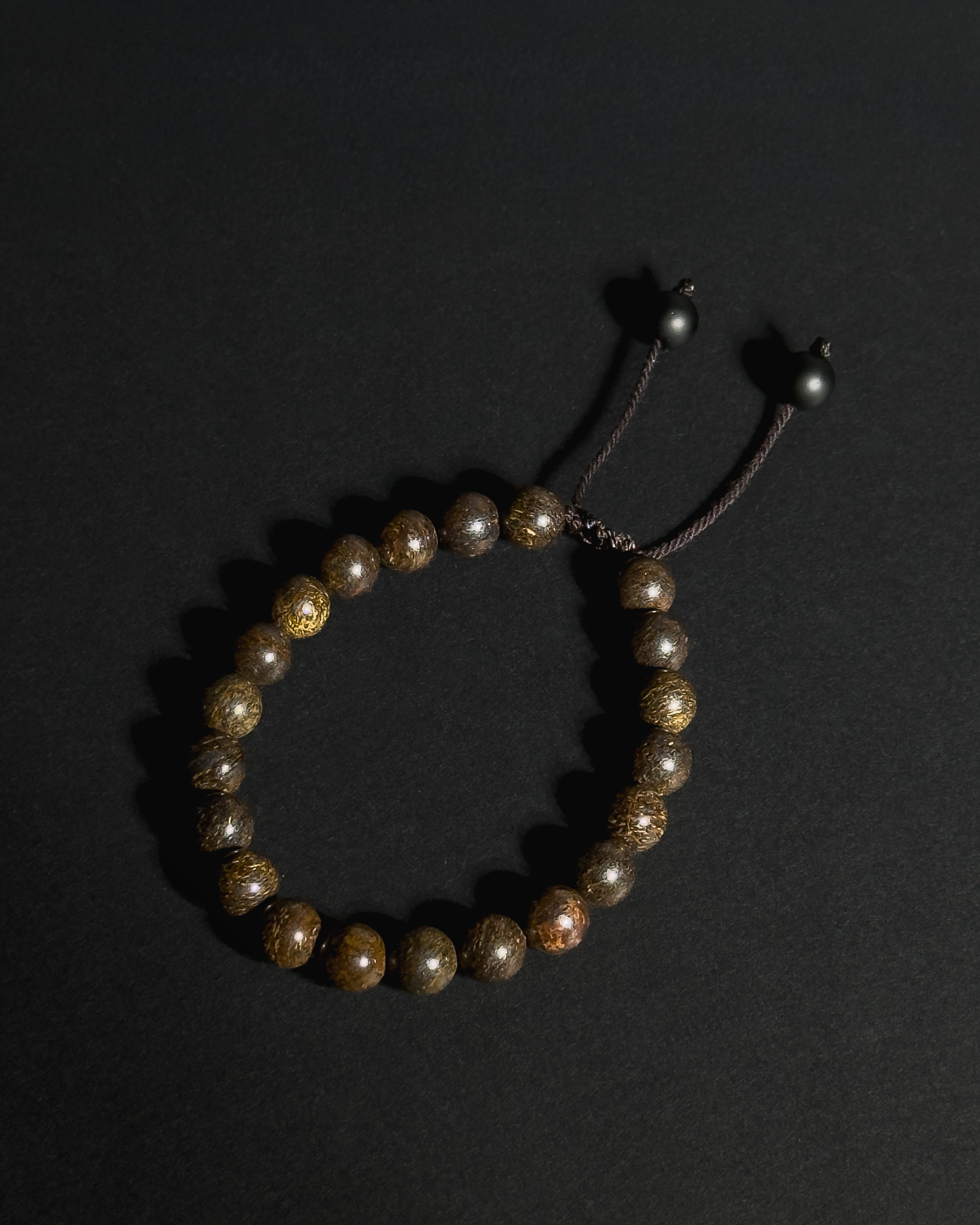 A beaded bracelet made of natural bronzite stone, featuring a dark adjustable cord closure with metal beads, displayed on a dark background.