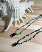 Green beaded necklace with black faceted crystal fish on a wooden surface next to a large shell.