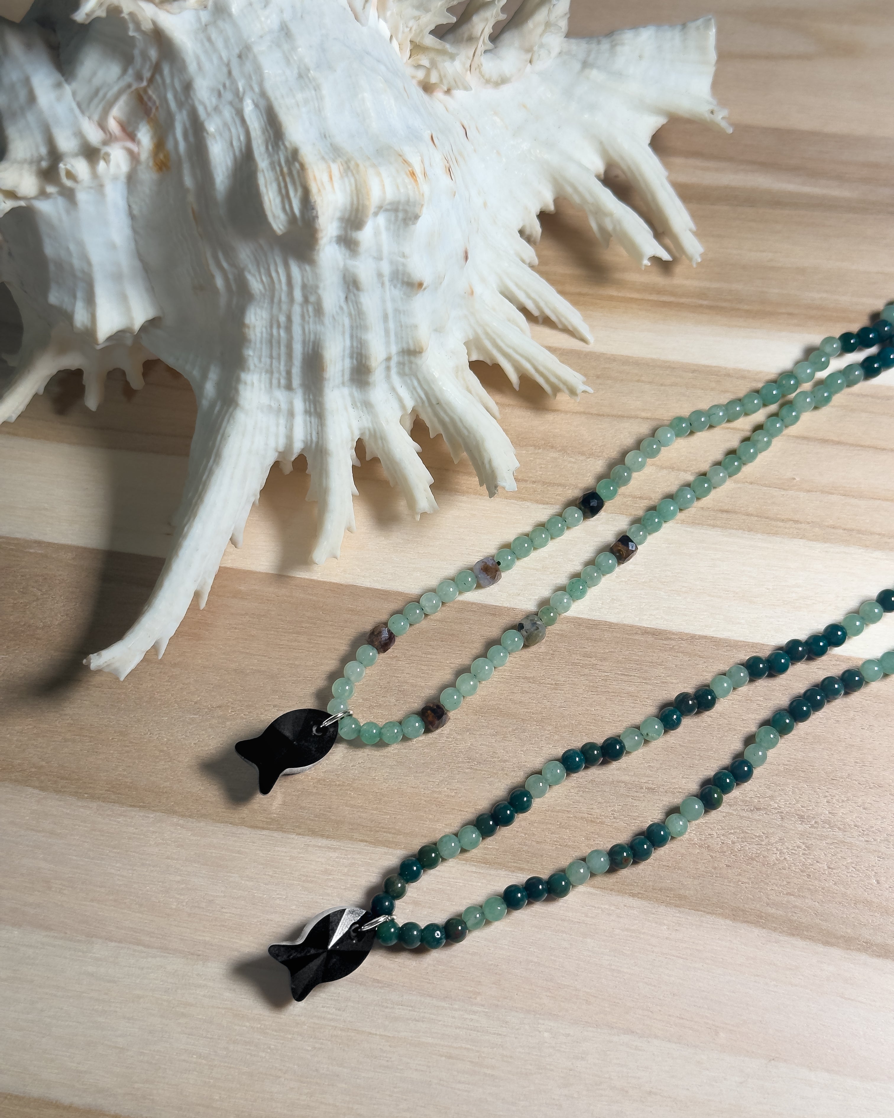 Green beaded necklace with black faceted crystal fish on a wooden surface next to a large shell.