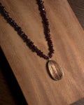 A beaded necklace with dark red garnet chips and a polished oval rutilated quartz pendant featuring natural wood grain, displayed on a warm brown wooden surface.