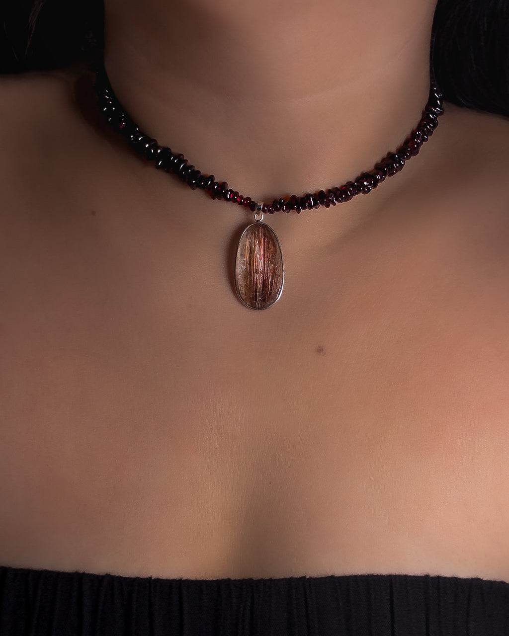 A close-up of a person's neck and chest wearing a beaded choker necklace with a central oval pendant. The necklace features dark red beads and a metallic copper-toned pendant with a silver border.