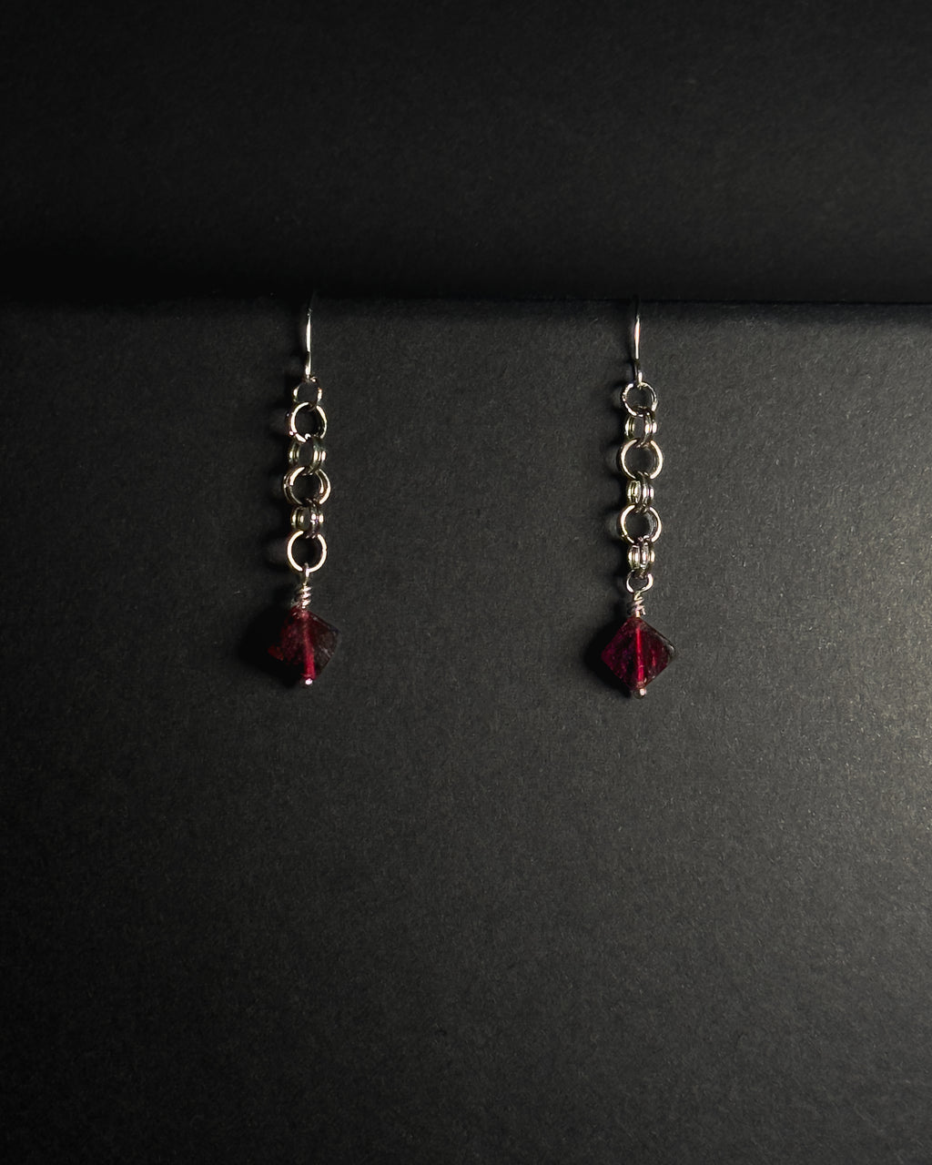 Stainless Steel hooked dangle earrings made with jumprings link by link with a geometric garnet against dark background.