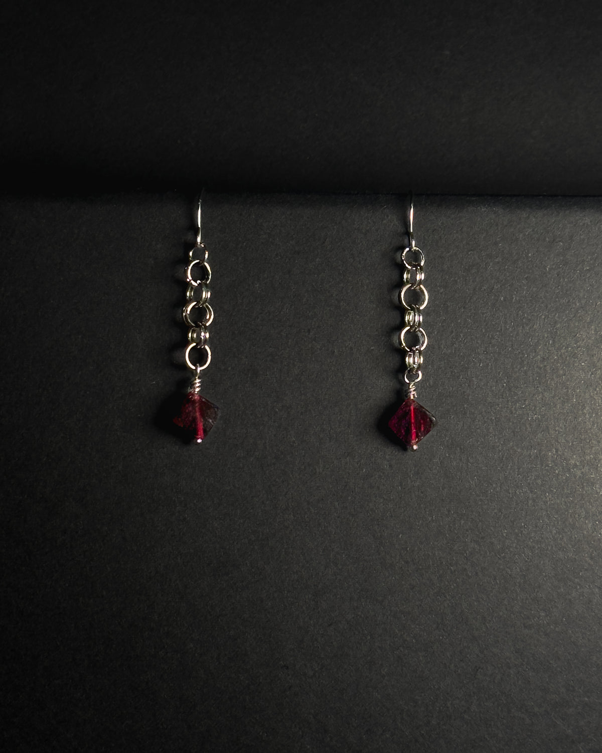Stainless Steel hooked dangle earrings made with jumprings link by link with a geometric garnet against dark background.