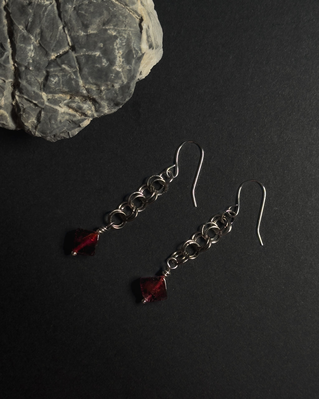 Stainless Steel hooked dangle earrings made with jumprings link by link with a geometric garnet against dark background with a natural rock.